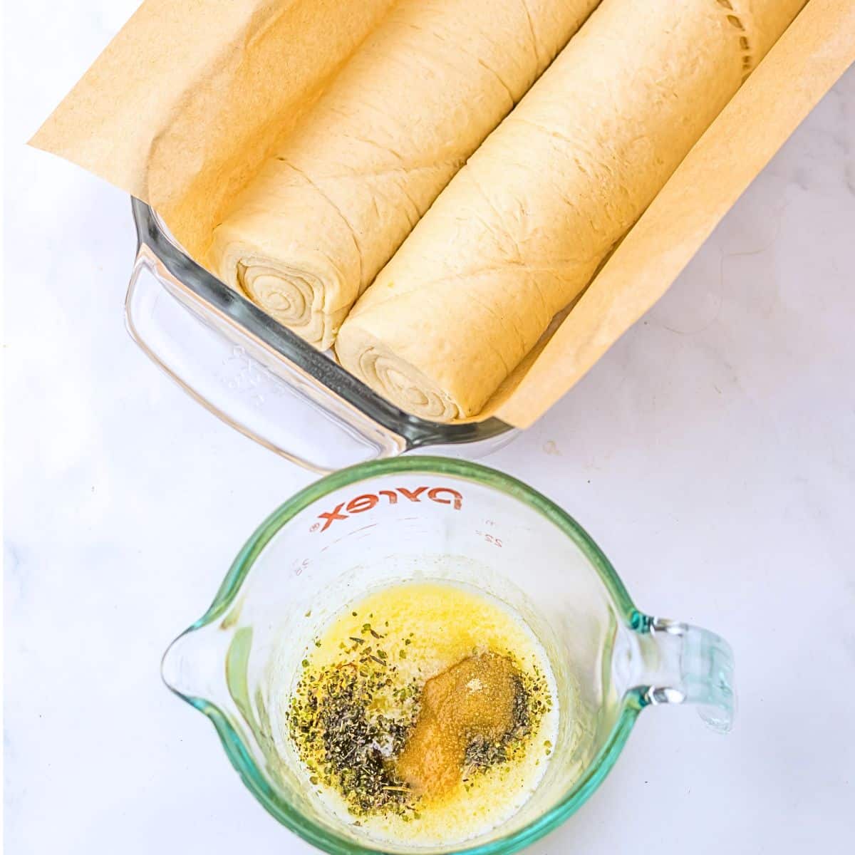 A glass loaf pan with the raw dough in it and a measuring cup in front of it with the melted butter and seasonings.